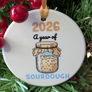 May include: A round white ceramic ornament with a gold ribbon, featuring the text 2026 A year of SOURDOUGH in tan, black, and light blue. The design includes a graphic of a glass jar filled with bubbly sourdough starter with a tan cloth lid.
