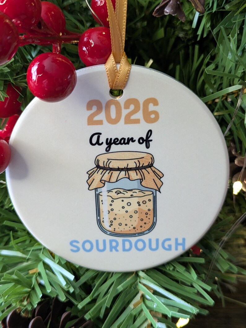 May include: A round white ceramic ornament with a gold ribbon, featuring the text 2026 A year of SOURDOUGH in tan, black, and light blue. The design includes a graphic of a glass jar filled with bubbly sourdough starter with a tan cloth lid.
