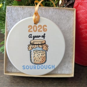 May include: A round white ceramic ornament featuring the text 2026 A year of SOURDOUGH and a graphic illustration of a glass jar filled with sourdough starter, topped with a tan cloth cover and secured with a gold ribbon for hanging.