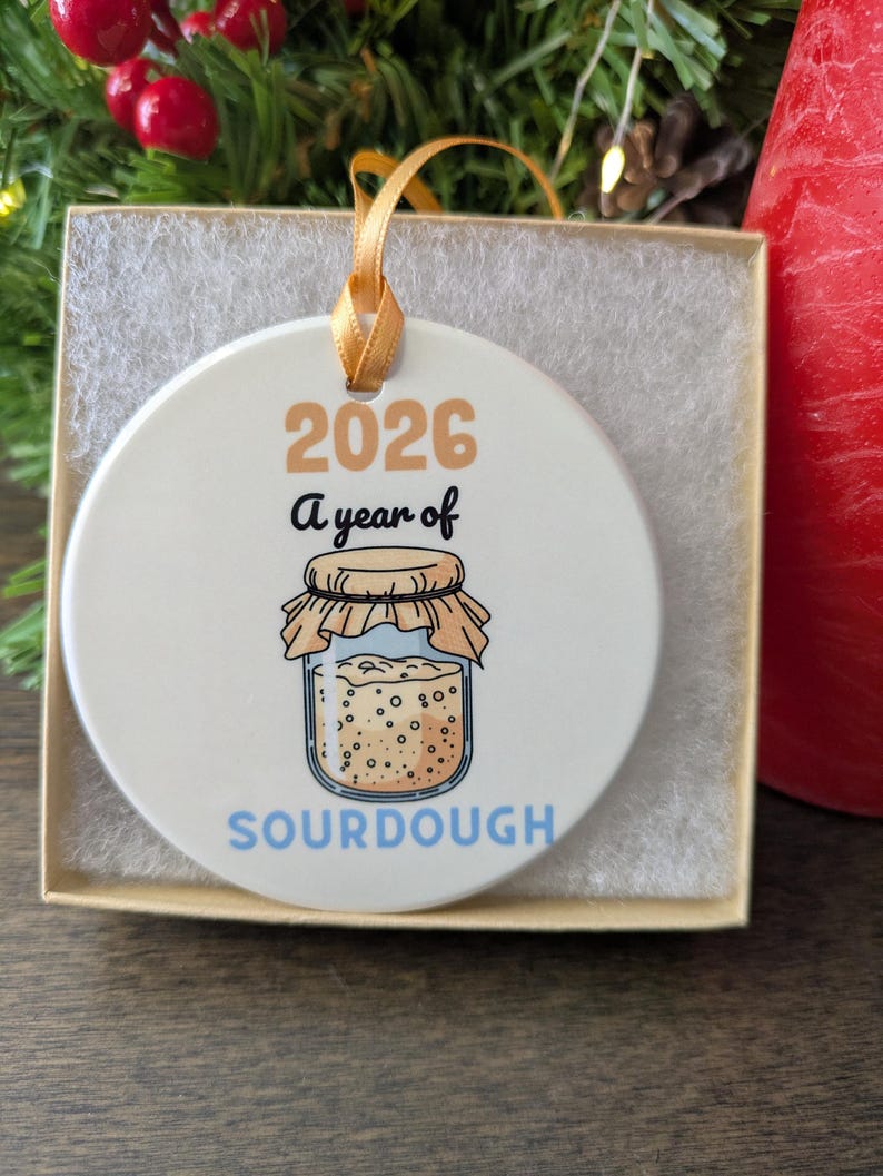May include: A round white ceramic ornament featuring the text 2026 A year of SOURDOUGH and a graphic illustration of a glass jar filled with sourdough starter, topped with a tan cloth cover and secured with a gold ribbon for hanging.