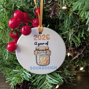 May include: A round white ceramic ornament hanging from a gold ribbon. The ornament features the text 2026 A year of SOURDOUGH and an illustration of a glass jar filled with sourdough starter, topped with a tan cloth cover and string.