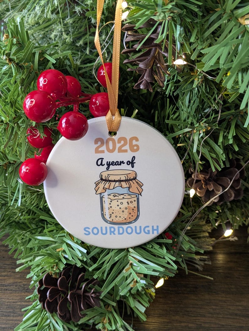 May include: A round white ceramic ornament hanging from a gold ribbon. The ornament features the text 2026 A year of SOURDOUGH and an illustration of a glass jar filled with sourdough starter, topped with a tan cloth cover and string.