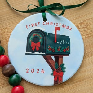 May include: A round white ceramic ornament with a green ribbon hanger. The design features a dark green mailbox with a red flag, a wreath, and the text "FIRST CHRISTMAS" and "1234 MAIN ST". The year "2026" is printed in red below the mailbox.