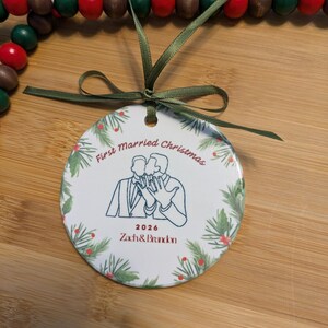 May include: A round white ceramic ornament with a green ribbon, featuring a line drawing of two people kissing and showing rings. Text reads First Married Christmas, 2026, and Zach & Brandon. The border has a green pine and red berry pattern.