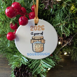 May include: A round white ceramic ornament hanging from a gold ribbon. The ornament features the text 2026 A year of SOURDOUGH in black, orange, and blue, with a central illustration of a glass jar filled with sourdough starter.