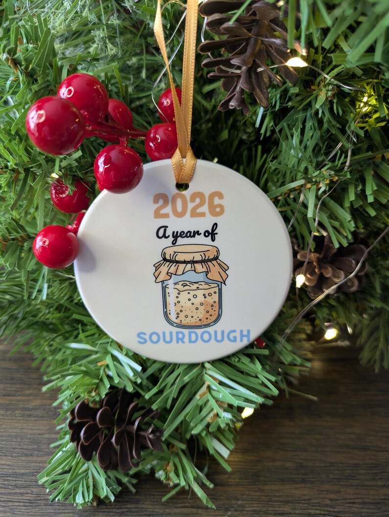 May include: A round white ceramic ornament hanging from a gold ribbon. The ornament features the text 2026 A year of SOURDOUGH in black, orange, and blue, with a central illustration of a glass jar filled with sourdough starter.