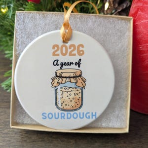 May include: A round white ceramic ornament featuring the text 2026 A year of SOURDOUGH in tan, black, and blue lettering. The design includes a graphic of a glass jar filled with bubbly sourdough starter, topped with a tan cloth cover.