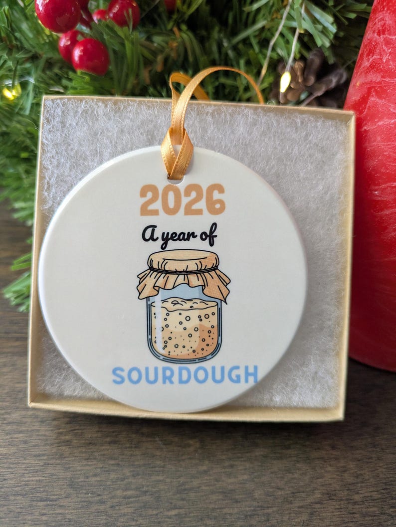 May include: A round white ceramic ornament featuring the text 2026 A year of SOURDOUGH in tan, black, and blue lettering. The design includes a graphic of a glass jar filled with bubbly sourdough starter, topped with a tan cloth cover.