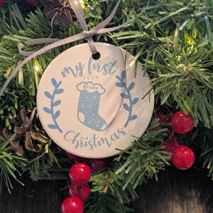May include: A round white ceramic ornament hanging by a grey ribbon. The ornament features blue text that reads My First Christmas 2026, a blue stocking graphic with the name Oliver printed on it, and two blue leafy branches on the sides.