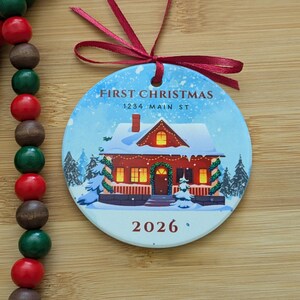 May include: A round ceramic Christmas ornament featuring a red house with snow on the roof, green garlands, and glowing windows. The text reads FIRST CHRISTMAS, 1234 MAIN ST, and 2026. A red ribbon is tied at the top, next to a string of wooden beads.