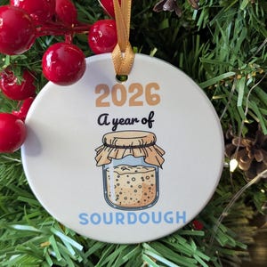 May include: A round white ceramic ornament hanging by a gold ribbon. The ornament features the text 2026 A year of SOURDOUGH in tan, black, and light blue, with a graphic illustration of a glass jar filled with sourdough starter.