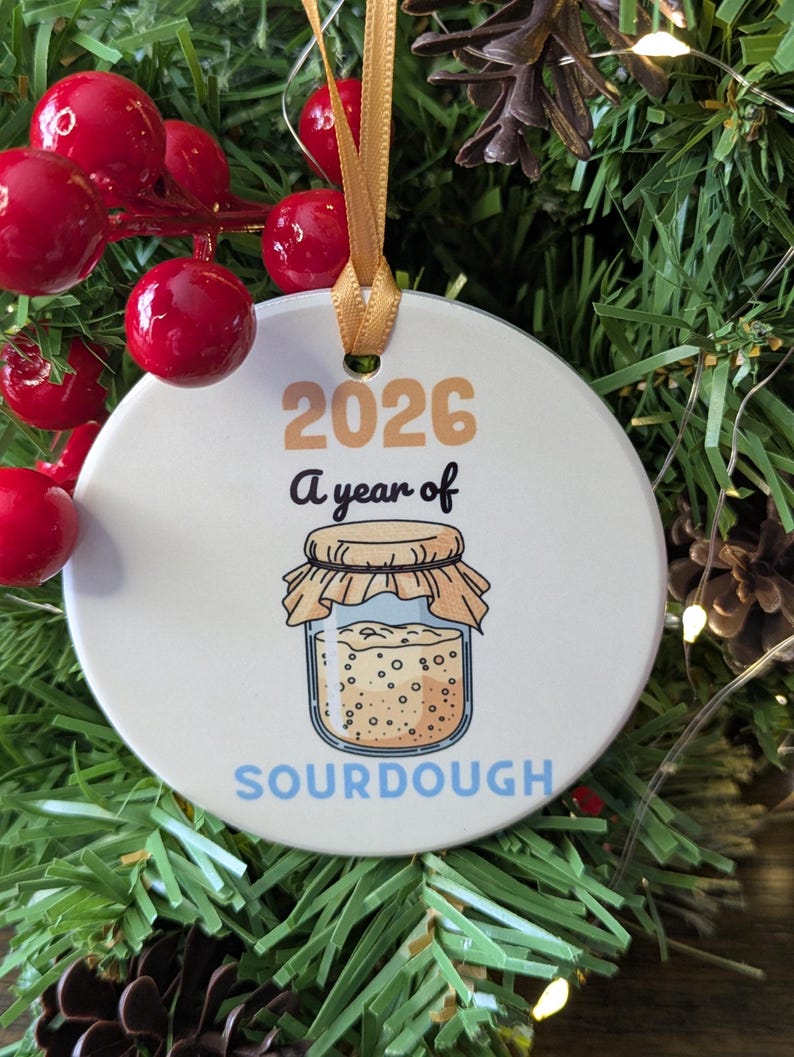 May include: A round white ceramic ornament hanging by a gold ribbon. The ornament features the text 2026 A year of SOURDOUGH in tan, black, and light blue, with a graphic illustration of a glass jar filled with sourdough starter.