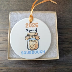 May include: A round white ceramic ornament with a gold ribbon hanger. The design features the text 2026, A year of, and SOURDOUGH in blue, with a graphic of a glass jar filled with bubbly sourdough starter covered by a cloth lid.
