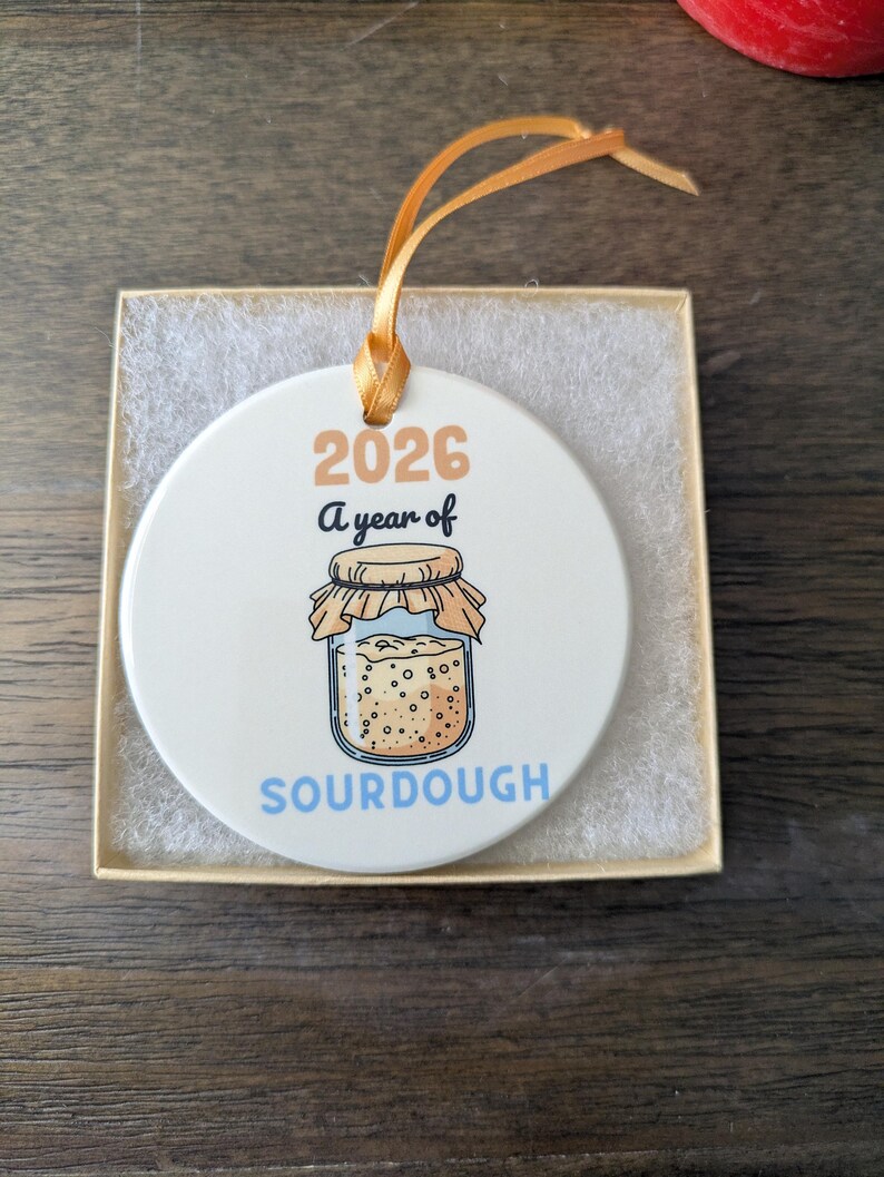 May include: A round white ceramic ornament with a gold ribbon hanger. The design features the text 2026, A year of, and SOURDOUGH in blue, with a graphic of a glass jar filled with bubbly sourdough starter covered by a cloth lid.