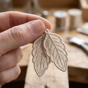 May include: Delicate, cream-colored feather earrings. The earrings are made of intricately woven lace, with a detailed pattern mimicking the structure of a feather. The earrings are held by a person's hand, showcasing the fine craftsmanship.