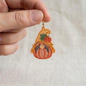 May include: An embroidered earring shaped like a pumpkin gnome with a gold-tone hook. The design features an orange pumpkin body, a tan hat with a small green leaf and pumpkin accent, and a dark gray mustache, all rendered in detailed thread work.