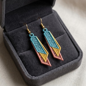 May include: A pair of dangle earrings featuring geometric embroidered shapes in teal, mustard yellow, and dusty pink thread. The earrings have gold-toned metal hooks and are presented in a dark gray velvet-lined jewelry box with a hinged lid.