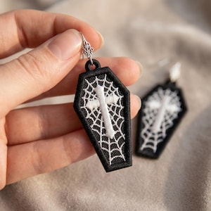 Puede incluir: Un par de pendientes colgantes con forma de ataúd, hechos de tela negra con patrones de telaraña bordados en blanco y una cruz blanca central. Los pendientes están sujetos a ganchos de metal plateado con detalles decorativos de filigrana.