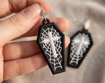 Puede incluir: Un par de pendientes colgantes con forma de ataúd, hechos de tela negra con patrones de telaraña bordados en blanco y una cruz blanca central. Los pendientes están sujetos a ganchos de metal plateado con detalles decorativos de filigrana.