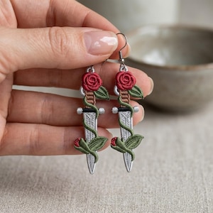 May include: A pair of drop earrings featuring embroidered dagger motifs. Each dagger has a silver-colored blade, a brown handle, and a red rose with green leaves wrapped around the blade. The earrings hang from silver-toned metal hooks.