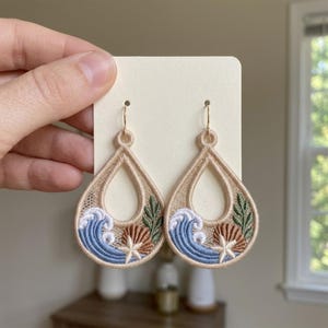 May include: A pair of teardrop-shaped embroidered earrings featuring a coastal design. The embroidery includes blue ocean waves, a white starfish, a brown seashell, and green seaweed on a beige lace background with gold-toned metal hooks.