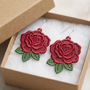 Könnte beinhalten: Ein Paar bestickte Ohrringe mit roten Rosen und grünen Blättern, präsentiert in einer offenen braunen Schachtel. Die Ohrringe haben silberfarbene Haken.