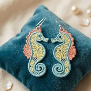 May include: A pair of seahorse earrings with intricate embroidery. The seahorses feature a light blue body, yellow interior details, and a coral-colored fin. The earrings are displayed on a teal velvet pillow.