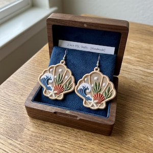 May include: A pair of 2 inch FSL shell earrings featuring intricate embroidery of a blue wave, green seaweed, a coral scallop shell, and a white starfish. The earrings are displayed in a dark wood box with a blue velvet interior and a label.