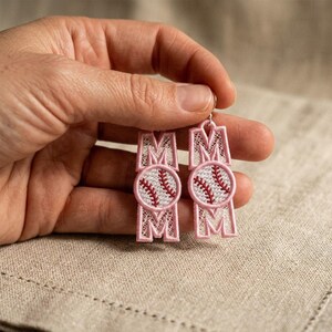 Puede incluir: Un par de pendientes colgantes con un diseño bordado que muestra la palabra MOM en hilo rosa claro. La letra O está sustituida por un gráfico de pelota de béisbol blanca con costuras rojas. El bordado tiene una textura delicada tipo encaje.