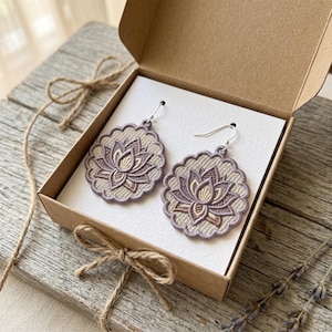 May include: A pair of round, embroidered earrings featuring a detailed lotus flower design in shades of mauve, tan, and cream. The earrings have a scalloped edge and silver-toned metal hooks, presented inside a square cardboard gift box.