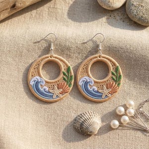 Puede incluir: Un par de pendientes circulares con un bordado intrincado de olas azules, una estrella de mar beige, una concha color melocotón y algas verdes sobre un fondo tostado, suspendidos de ganchos de metal plateado.