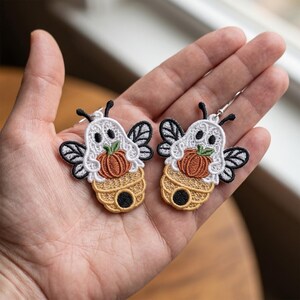 May include: A pair of embroidered earrings featuring a white ghost bee design. Each earring depicts a ghost with black eyes, holding an orange pumpkin, positioned above a tan beehive with a black entrance, and black and white wings.
