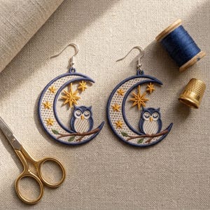 Könnte beinhalten: Ein Paar halbmondförmige Ohrringe mit gestickten Eulen auf Ästen und gelben Sternen. Die Ohrringe haben silberfarbene Haken und sind neben einer Spule mit blauem Garn, einem goldenen Fingerhut und einer goldenen Schere abgebildet.