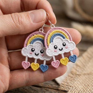 May include: A pair of embroidered dangle earrings featuring a white cloud with a smiling face, pink cheeks, and a rainbow arch above. Three small hearts in pink, yellow, and blue hang from the bottom of each cloud on silver-toned metal hooks.