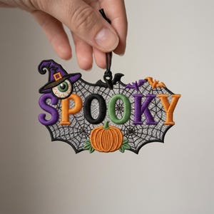 May include: An embroidered hanging ornament featuring a spiderweb background with the word SPOOKY in multi-colored letters. It includes a witch hat with an eyeball, a small orange pumpkin, and two small bats, all finished with a black cord.