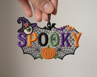 Puede incluir: Un adorno colgante bordado con un fondo de telaraña y la palabra SPOOKY en letras multicolores. Incluye un sombrero de bruja con un globo ocular, una pequeña calabaza naranja y dos pequeños murciélagos, todo con un cordón negro.