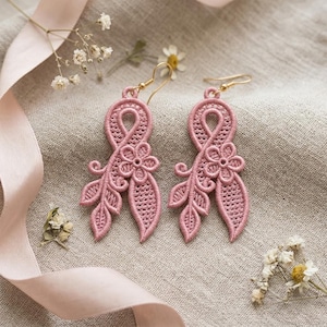 Puede incluir: Un par de pendientes de encaje en color rosa empolvado con un intrincado diseño bordado de una cinta en bucle, una pequeña flor y dos hojas puntiagudas. Los pendientes cuelgan de ganchos de metal dorado y muestran una textura calada detallada.
