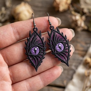 May include: A pair of drop earrings featuring an embroidered eye design. The eye has a vibrant purple iris with a white highlight, surrounded by intricate black thread detailing that forms a pointed, leaf-like shape. Dark metal hooks attach at the top.