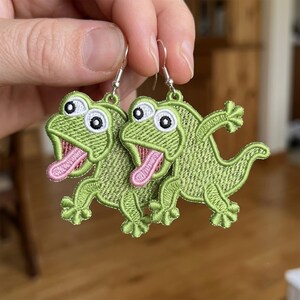 May include: A pair of dangle earrings featuring embroidered green lizard figures with large white eyes, black pupils, and protruding pink tongues. The lizards are crafted with detailed green thread stitching and are attached to silver-toned metal hooks.