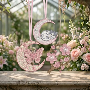 May include: Three hanging ornaments featuring intricate embroidery. One is a crescent moon with a sleeping cat, one is a crescent moon with a butterfly, and one is a circular frame with pink cherry blossoms, all suspended by ribbons.