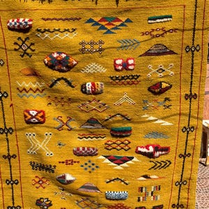 May include: Mustard yellow woven rug with a geometric pattern. The rug features various shapes and designs in red, white, blue, and green. The rug has a fringe at the top and a black border with red accents.