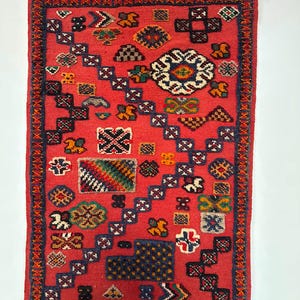 May include: A rectangular red rug featuring a central diagonal band of dark blue geometric shapes. The surface is decorated with various multicoloured motifs, including floral patterns, squares, and crosses. The rug has white fringe on both ends.