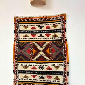 May include: A rectangular woven rug featuring a central geometric diamond pattern in shades of red, orange, and dark grey. The design includes horizontal bands with small floral motifs in red, blue, and green, finished with white fringe at both ends.