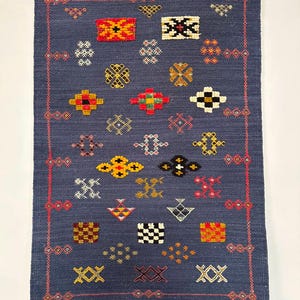May include: A rectangular navy blue woven rug featuring a variety of small, colourful embroidered geometric patterns in red, yellow, white, and black. The rug is framed by a thin red border and finished with white fringe along the top and bottom edges.