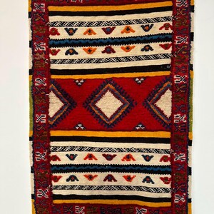 May include: A rectangular woven rug featuring a central red panel with three diamond shapes, bordered by horizontal stripes in cream, black, and yellow. The design includes small geometric motifs in blue, orange, and red, with white fringe at both ends.