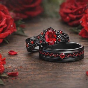 Puede incluir: Dos anillos negros con piedras preciosas rojas, sobre un fondo de rosas rojas. Un anillo presenta una gran gema ovalada, mientras que el otro tiene una banda roja decorativa. Los anillos se exhiben sobre una superficie de madera.
