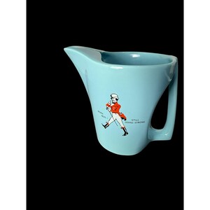 May include: A light blue ceramic pitcher featuring a graphic of a man in a red coat and top hat walking with a cane. The text BORN 1820 and STILL GOING STRONG is printed on the side of the pitcher, which has a wide spout and a sturdy handle.