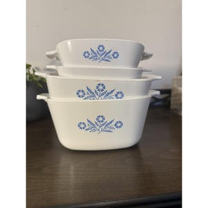 May include: Four white casserole dishes with blue floral patterns are stacked. Each dish has a rectangular shape with rounded edges and handles. The blue floral design is centered on the front of each dish. The dishes are stacked on a dark wooden surface.