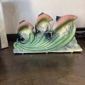 May include: A ceramic figurine featuring three stylized fish leaping through sculpted waves. The piece is finished in a glossy glaze with blended shades of sage green, deep blue, and soft pink, creating a fluid, textured appearance.