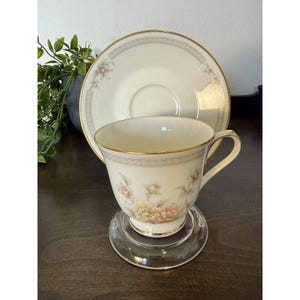 Peut inclure: Un service à thé crème avec un motif floral. La tasse et la soucoupe ont une bordure dorée et un motif délicat de fleurs roses, jaunes et vertes. La tasse est posée sur un dessous de verre rond et transparent.